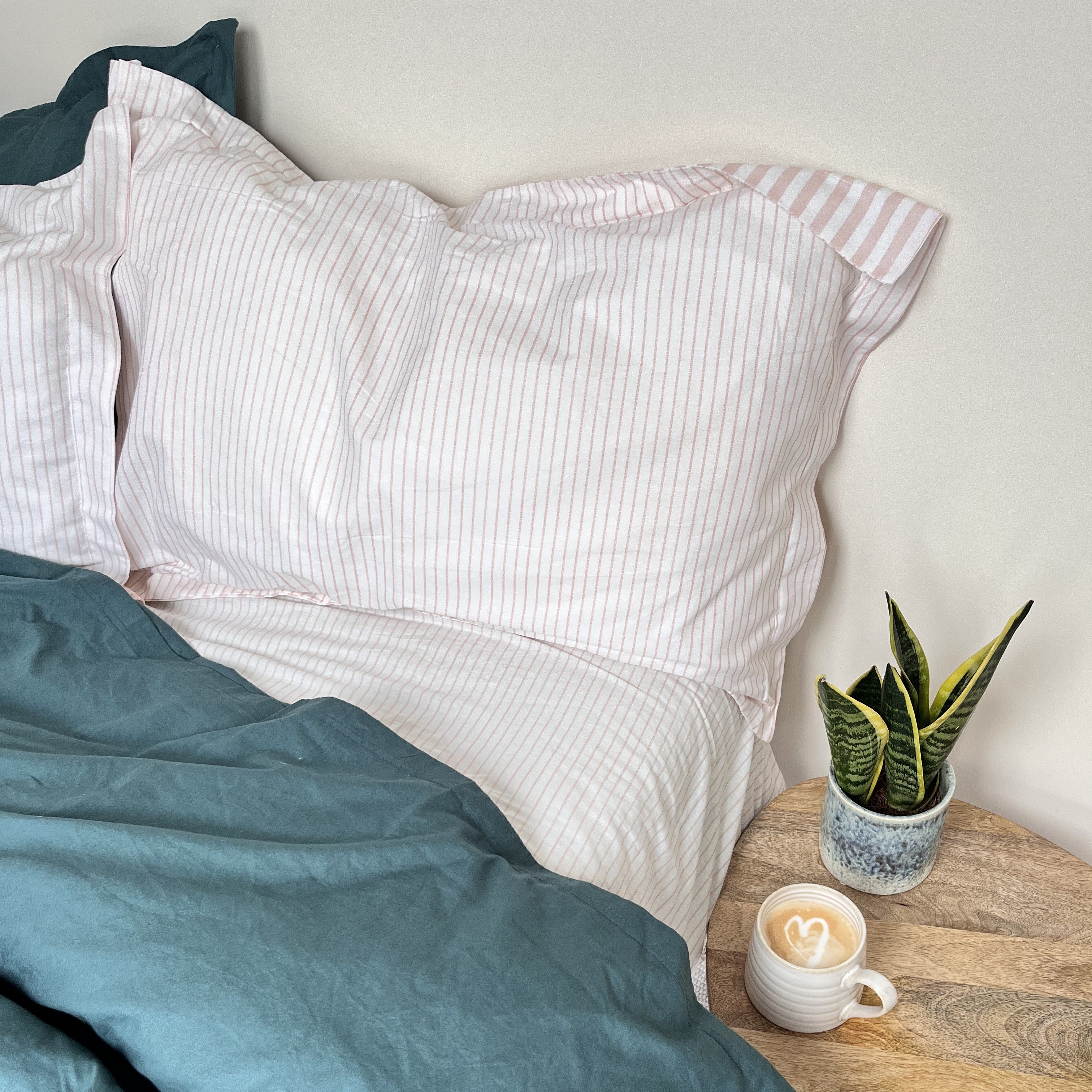 Pink striped pillowcases with dark teal duvet on a bed with a snake plant and coffee cup on the table