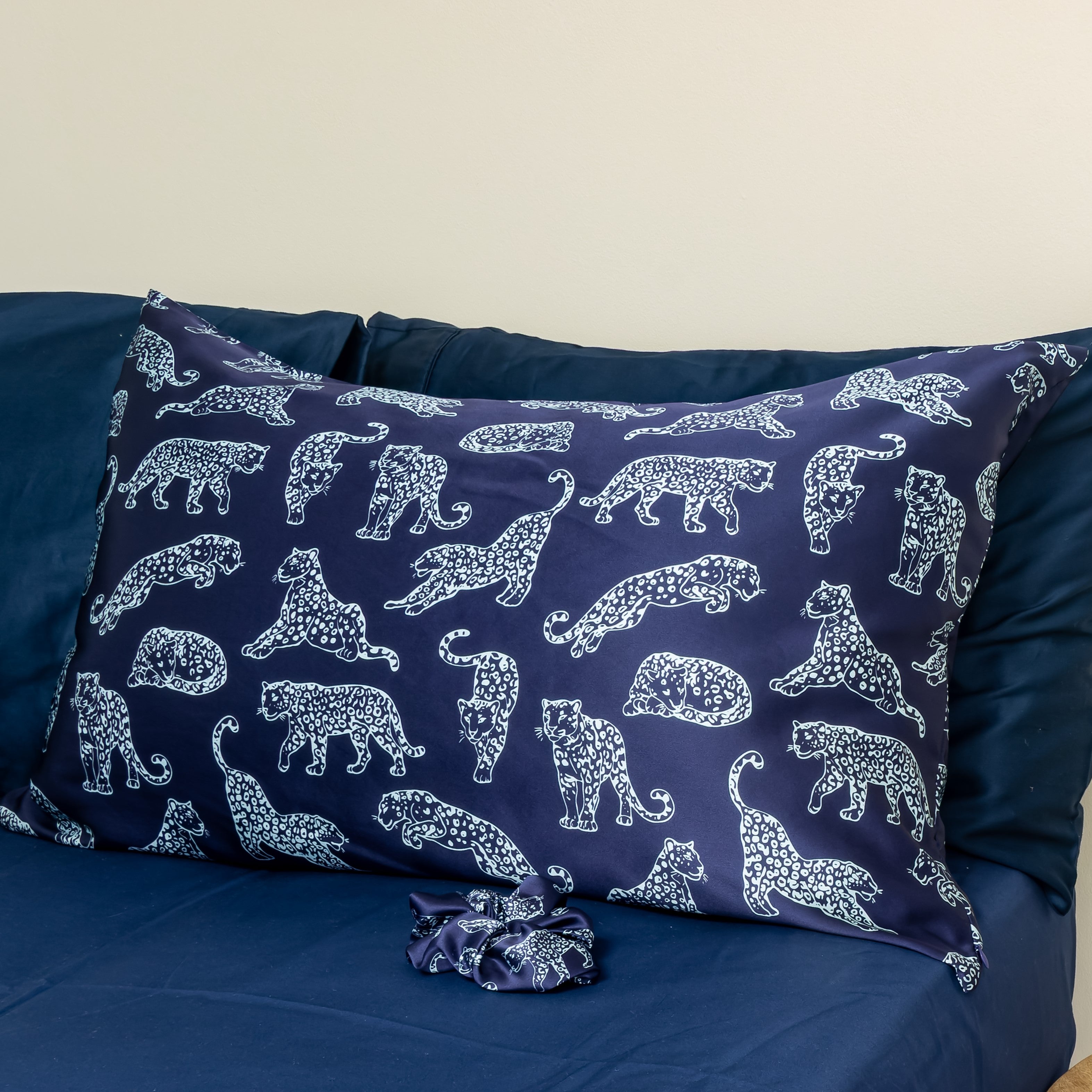 Lifestyle photo of a MYRN Silk navy blue leopard print silk pillowcase with a matching silk scrunchie on a blue bed. 