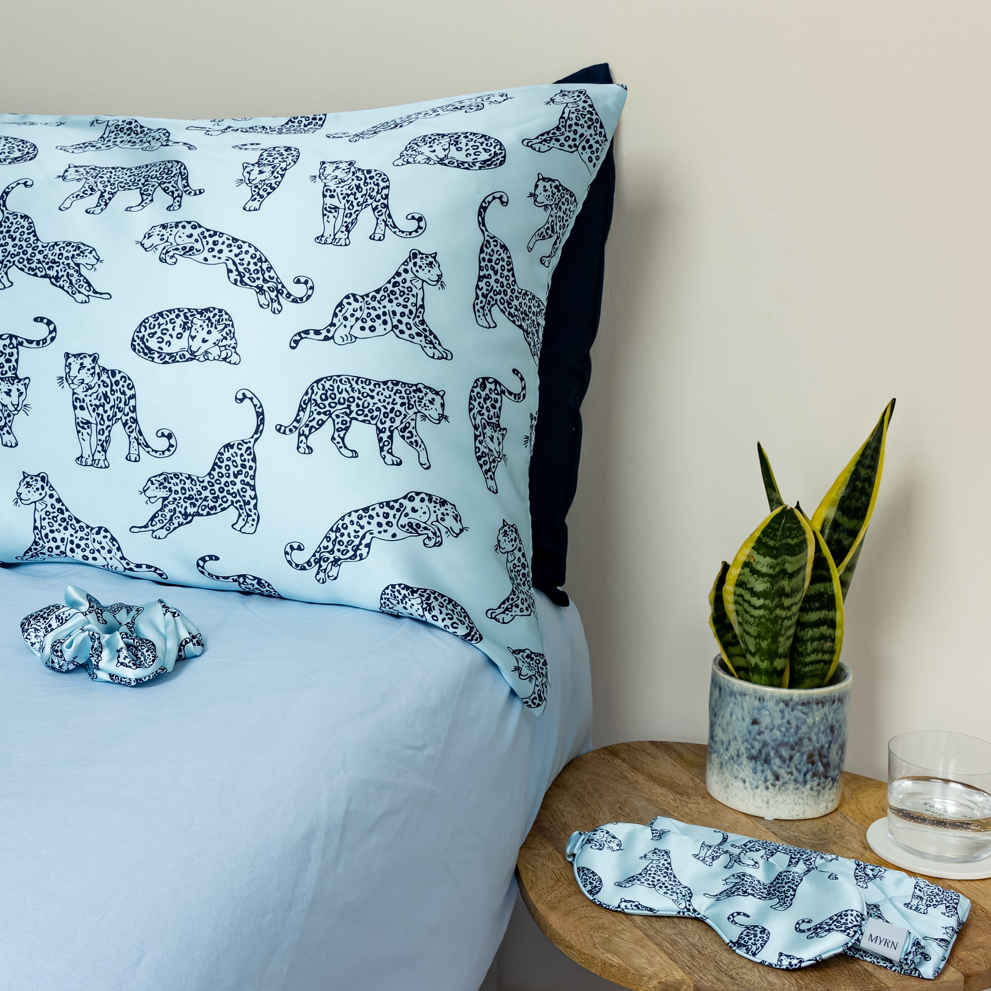 Light blue MYRN Silk leopard print pillowcase and silk scrunchie on a blue bed, with a matching eye mask and travel case on the side table.