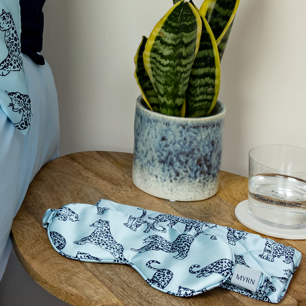 Light blue leopard print MYRN Silk eye mask and travel case on a bedside table next to a snake plant.