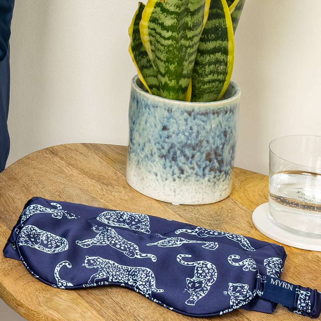 Lifestyle photo of a MYRN Silk navy blue leopard print silk eye mask with a matching silk travel on a wooden side table, with a snake plant and a glass of water.