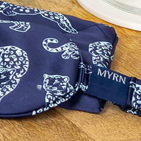 Close up lifestyle photo of half a MYRN Silk navy blue leopard print silk eye mask with a matching silk travel on a wooden side table. The eye mask logo is clearly in view.