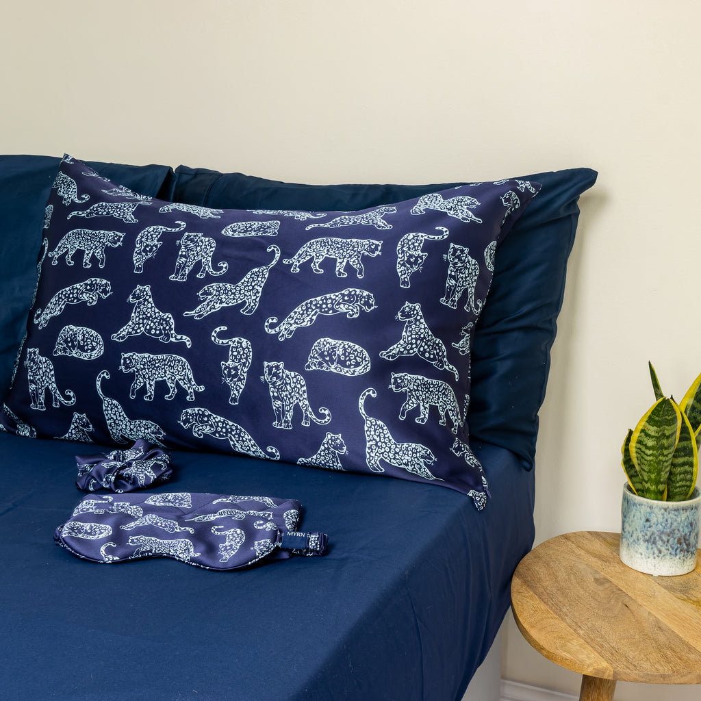 MYRN Silk Navy Blue Leopard print silk pillowcase, eye mask, travel case and silk scrunchie on a bed.