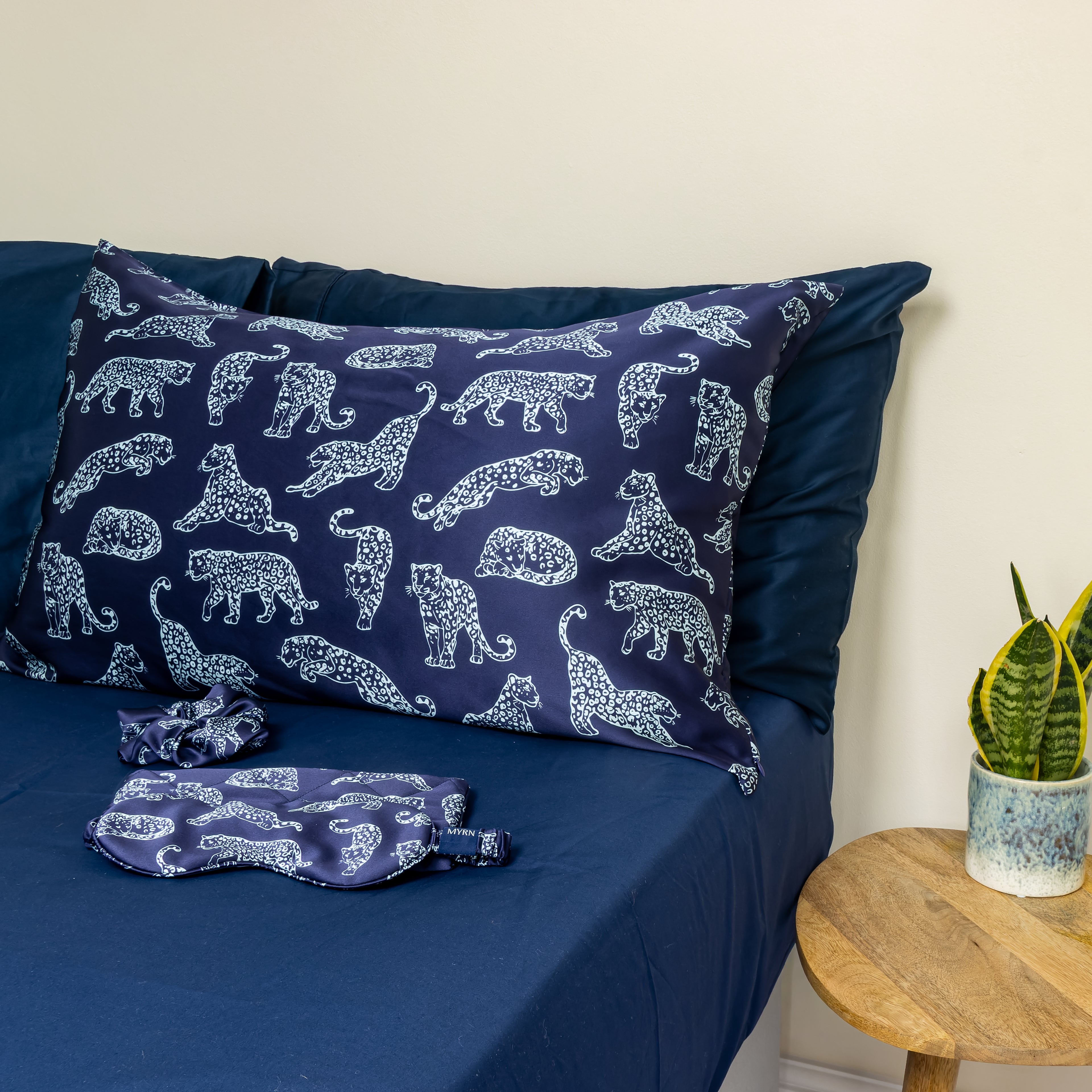 MYRN Silk Navy Blue Leopard print silk pillowcase, eye mask, travel case and silk scrunchie on a bed.