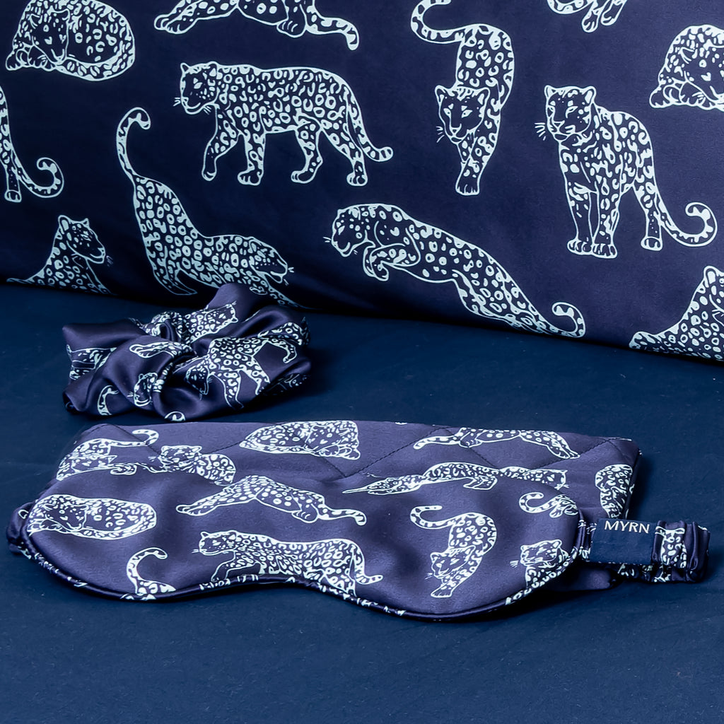 Close up of a dark blue MYRN Silk leopard print eye mask, travel case and scrunchie o a bed with the edge of a silk pillowcase appearing.
