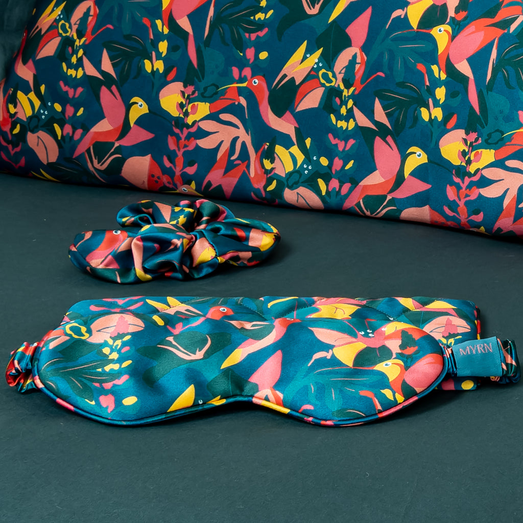 Close up shot of a MYRN Silk hummingbird eye mask on a green bedding set with a matching silk scrunchie and silk pillowcase.