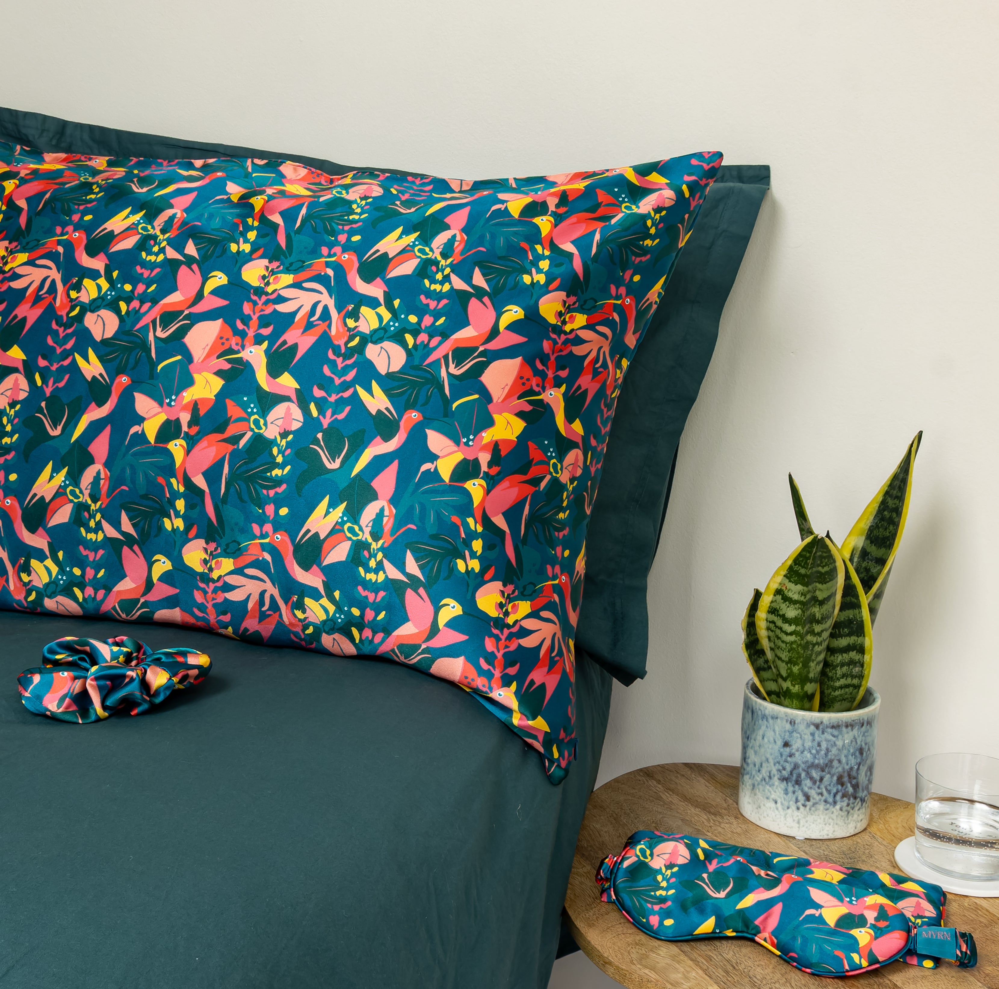 MYRN Silk Hummingbird printed silk pillowcase on a green bed with a matching silk scrunchie. The beside table holds a matching eye mask and travel case.