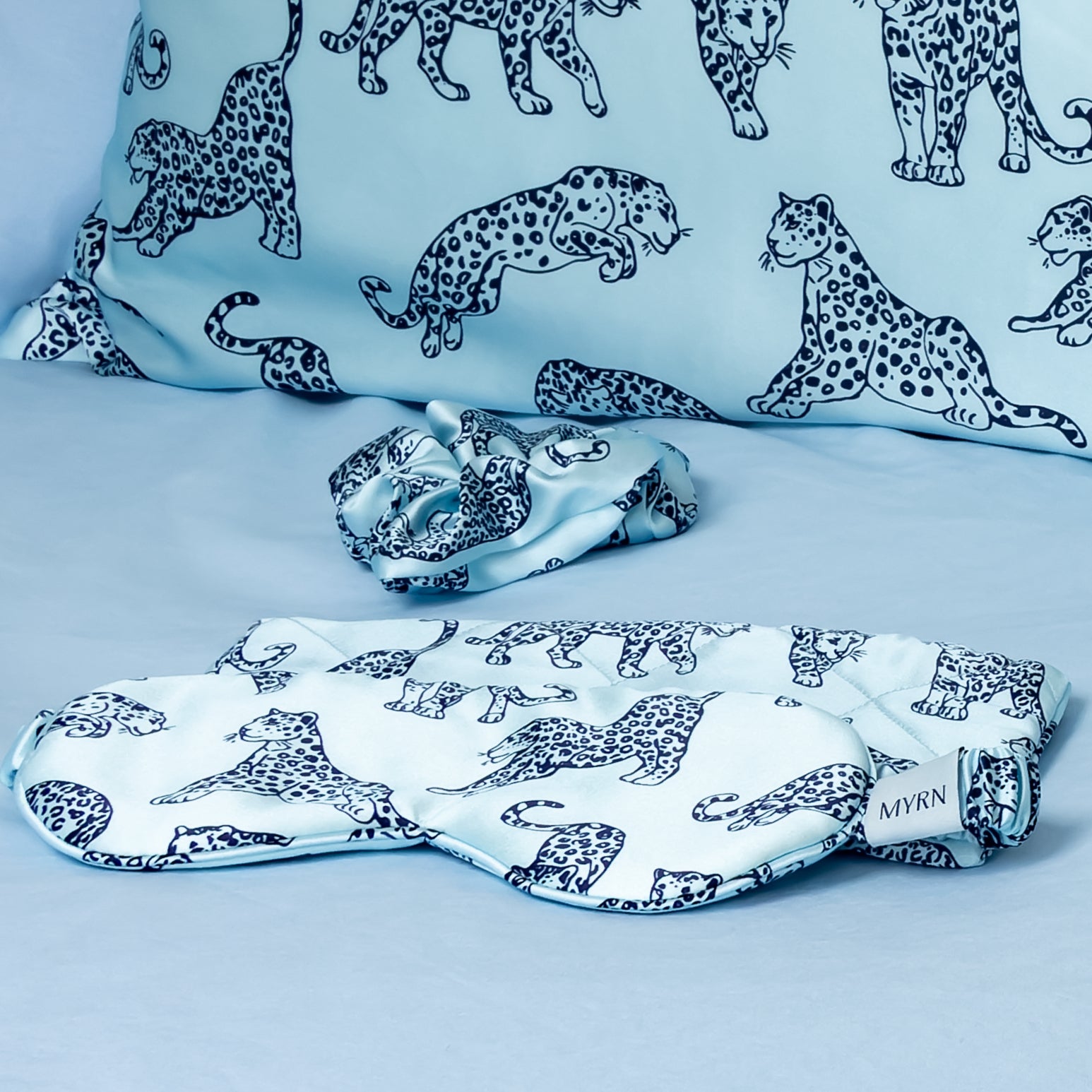 Close up of a MYRN Silk light blue leopard print eye mask, travel case and scrunchie next to a silk pillowcase.