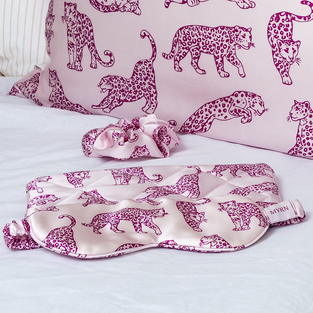 Close up of a MYRN Silk pink leopard print eye mask and travel case, as well as silk scrunchie. Laying on a bed next t the bottom of a pink leopard print silk pillowcase.