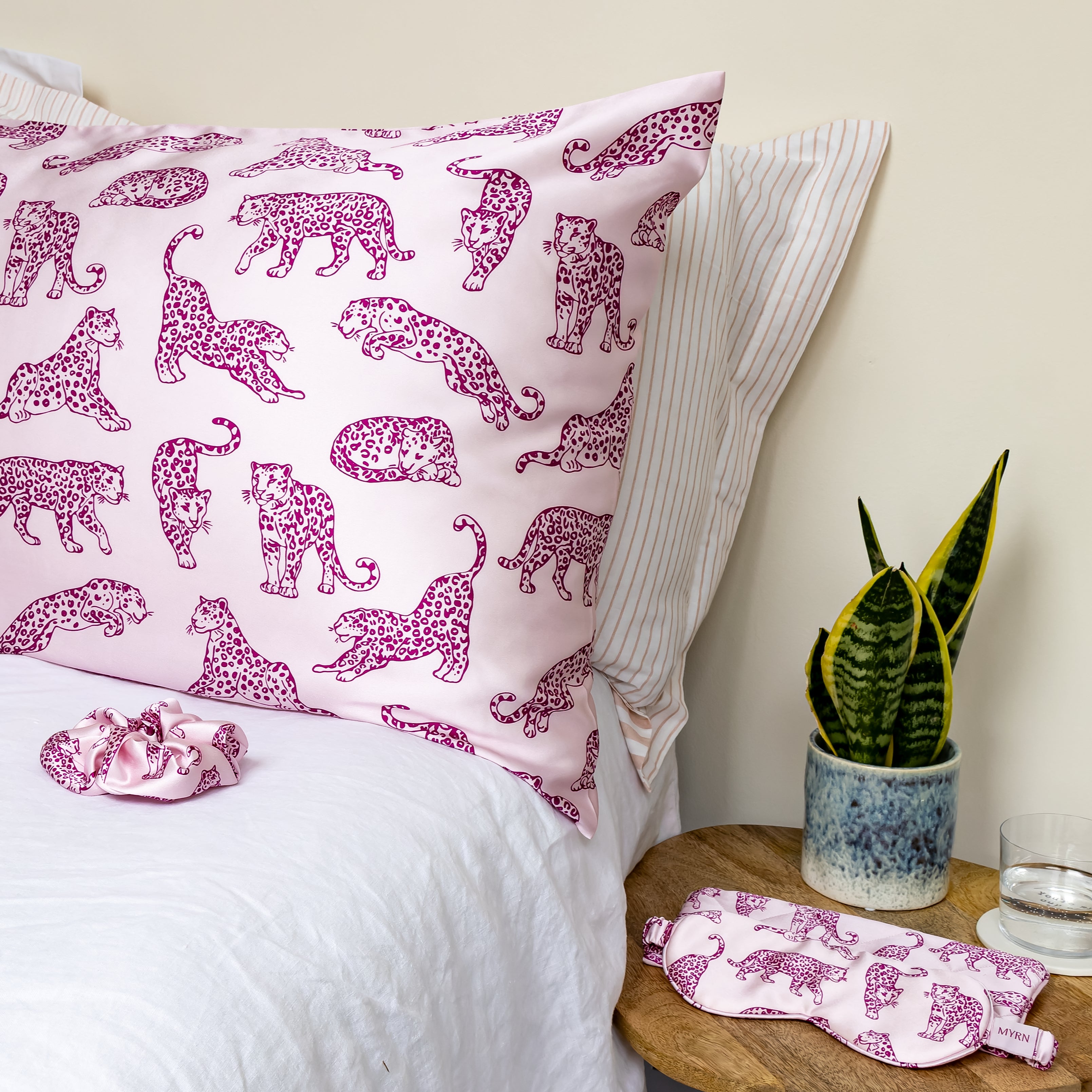 MYRN Pink leopard print silk pillowcase on a bed with a silk scrunchie. A matching silk eye mask is on the table next to the bed.