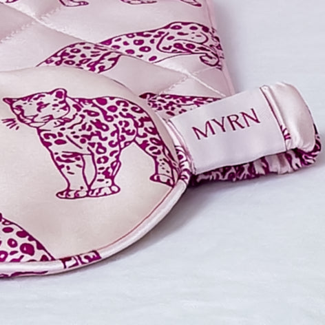 Close up of half a MYRN Silk Pink leopard print silk eye mask and matching travel case and logo label on a bed.