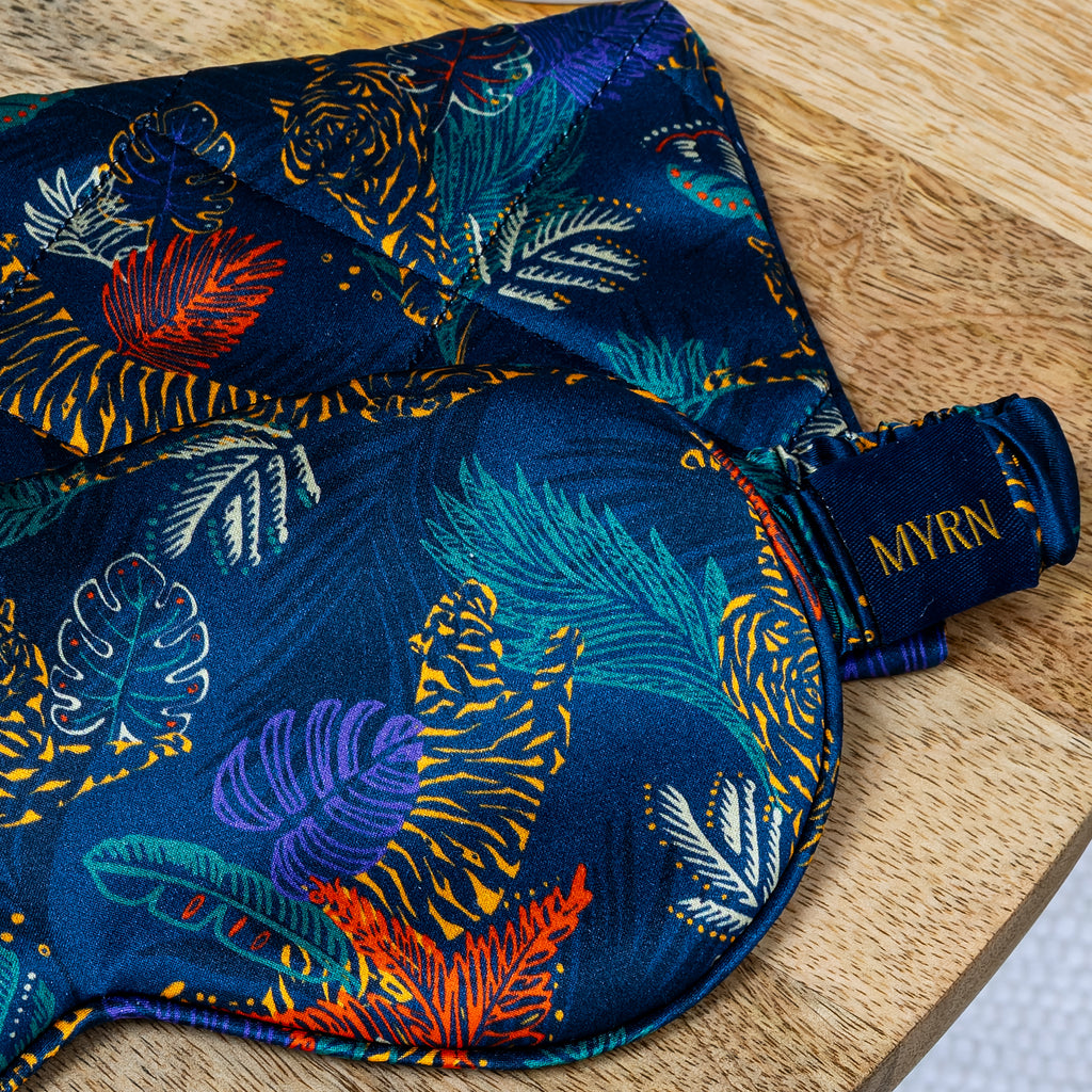 Close up lifestyle photo of half a MYRN Silk tiger print silk eye mask with a matching silk travel on a wooden side table. The eye mask logo is clearly in view.