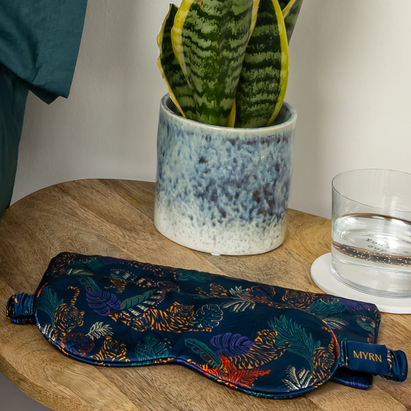 Lifestyle photo of a MYRN Silk navy blue tiger print silk eye mask with a matching silk travel on a wooden side table, with a snake plant and a glass of water.