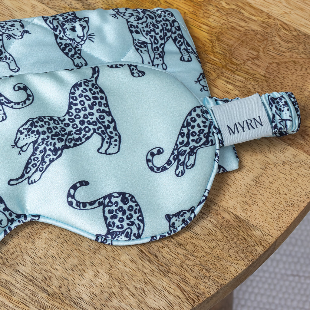 Close up of half of a light blue MYRN Silk leopard print eye mask and quilted travel case on a bedside table.