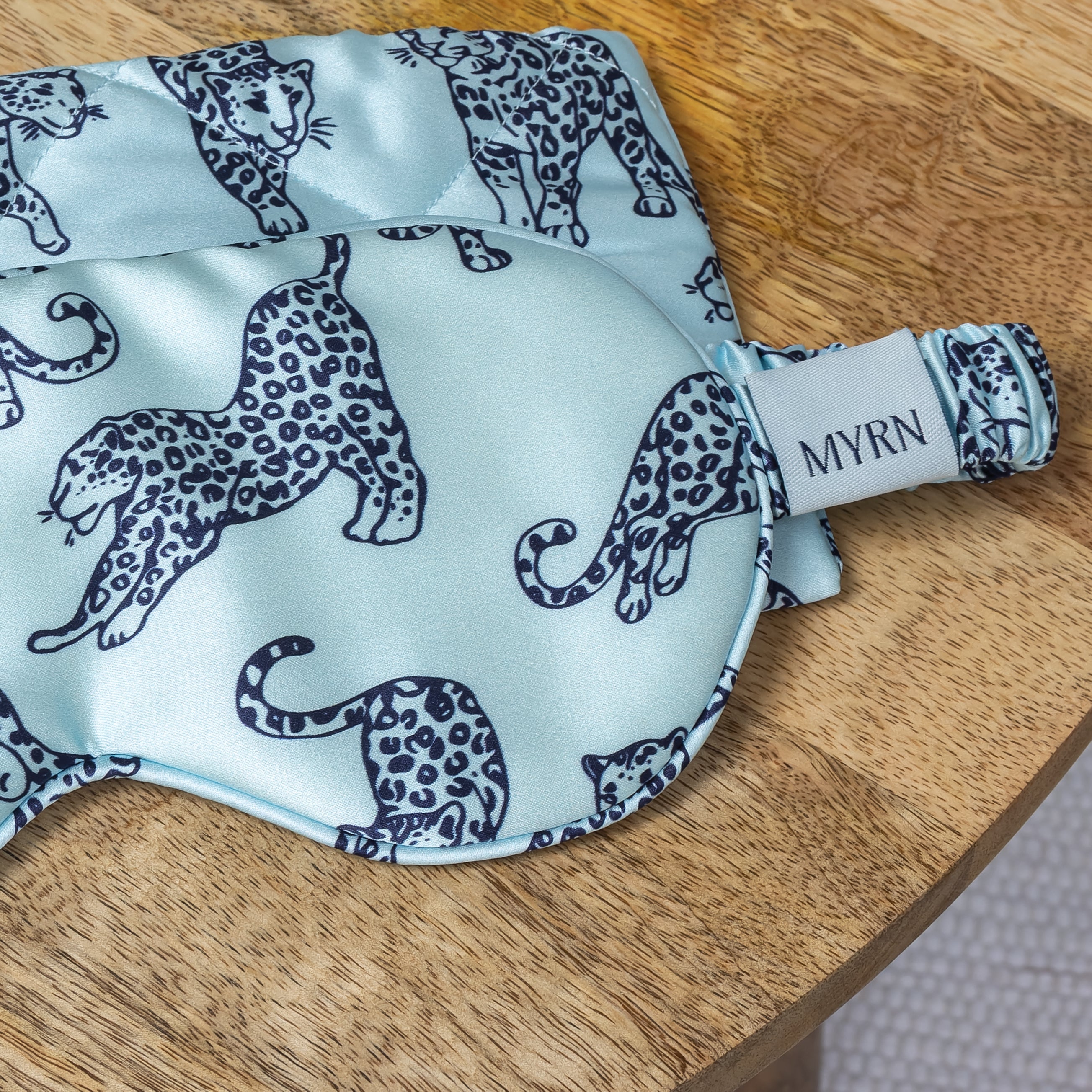 Close up of half of a light blue MYRN Silk leopard print eye mask and quilted travel case on a bedside table.