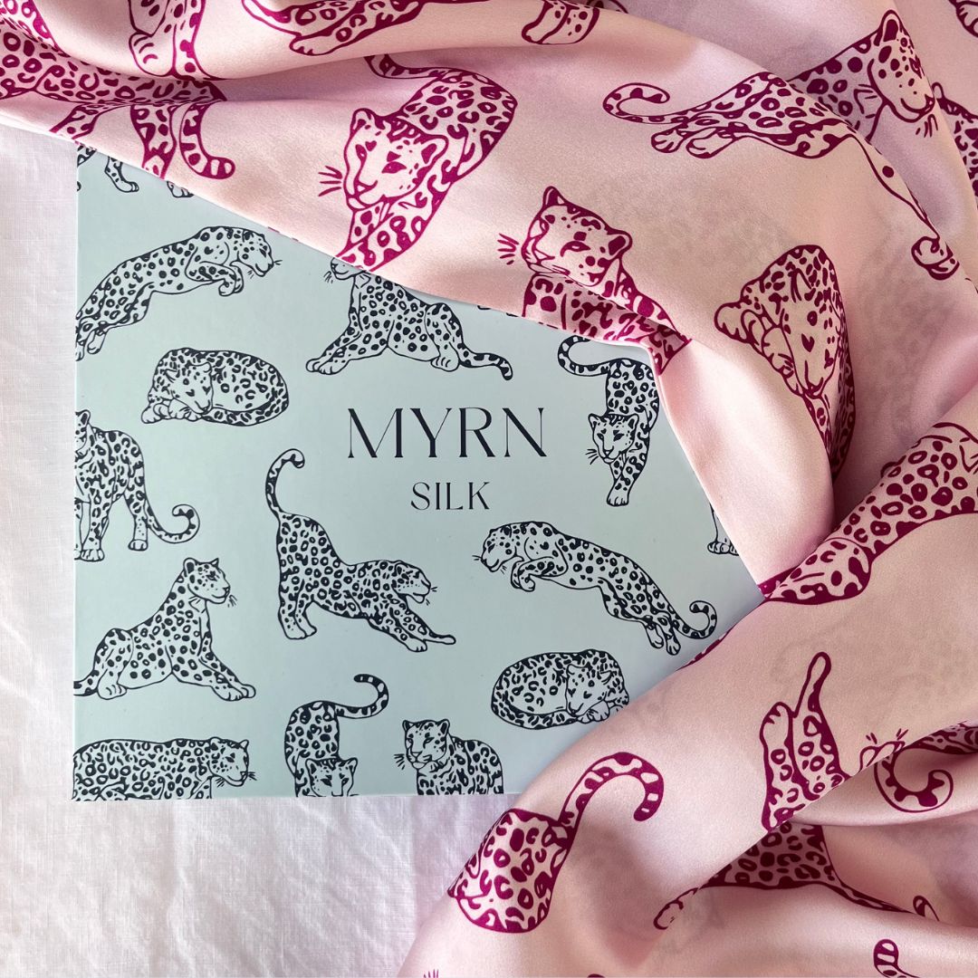 MYRN Silk pink leopard print pillowcase draped over a MYRN Silk branded gift box laying on a light pink bed.