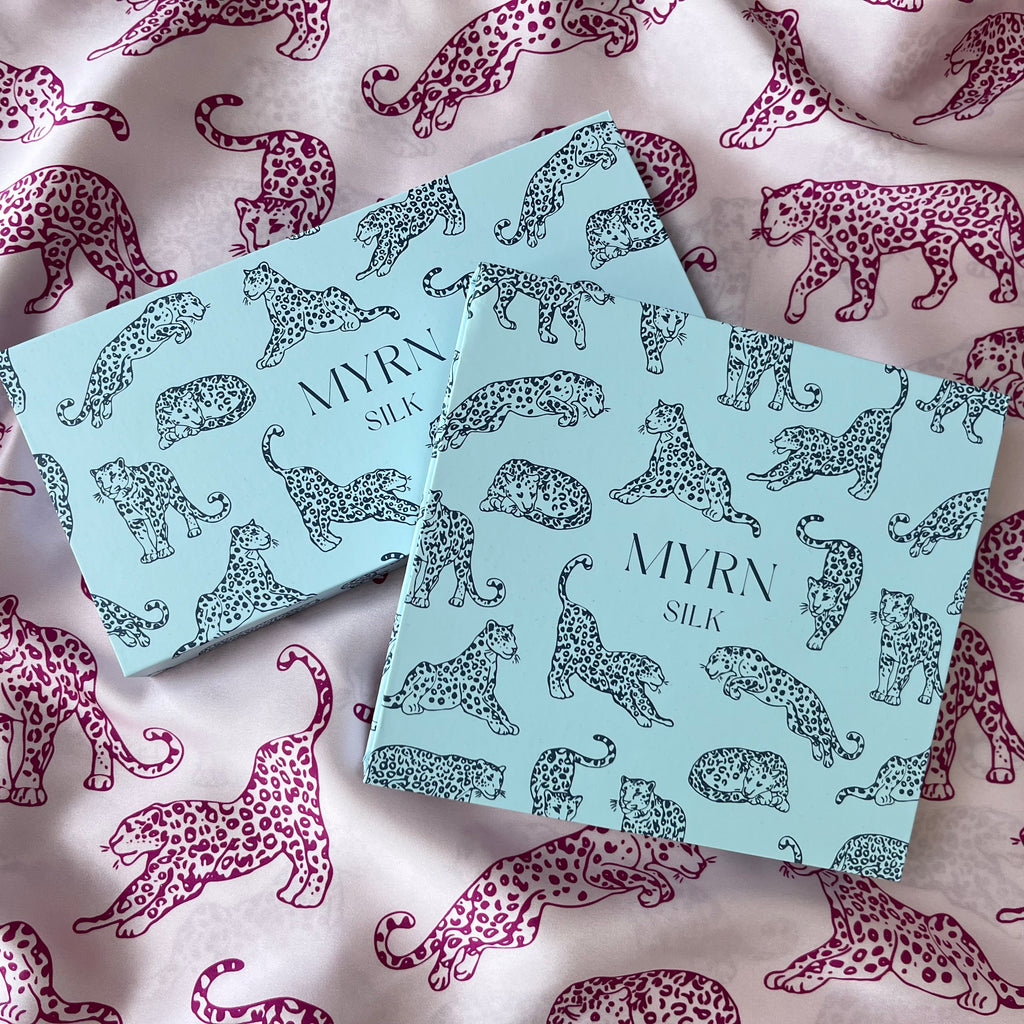 Two MYRN Silk branded presentation boxes laying on draped pink leopard print fabric.