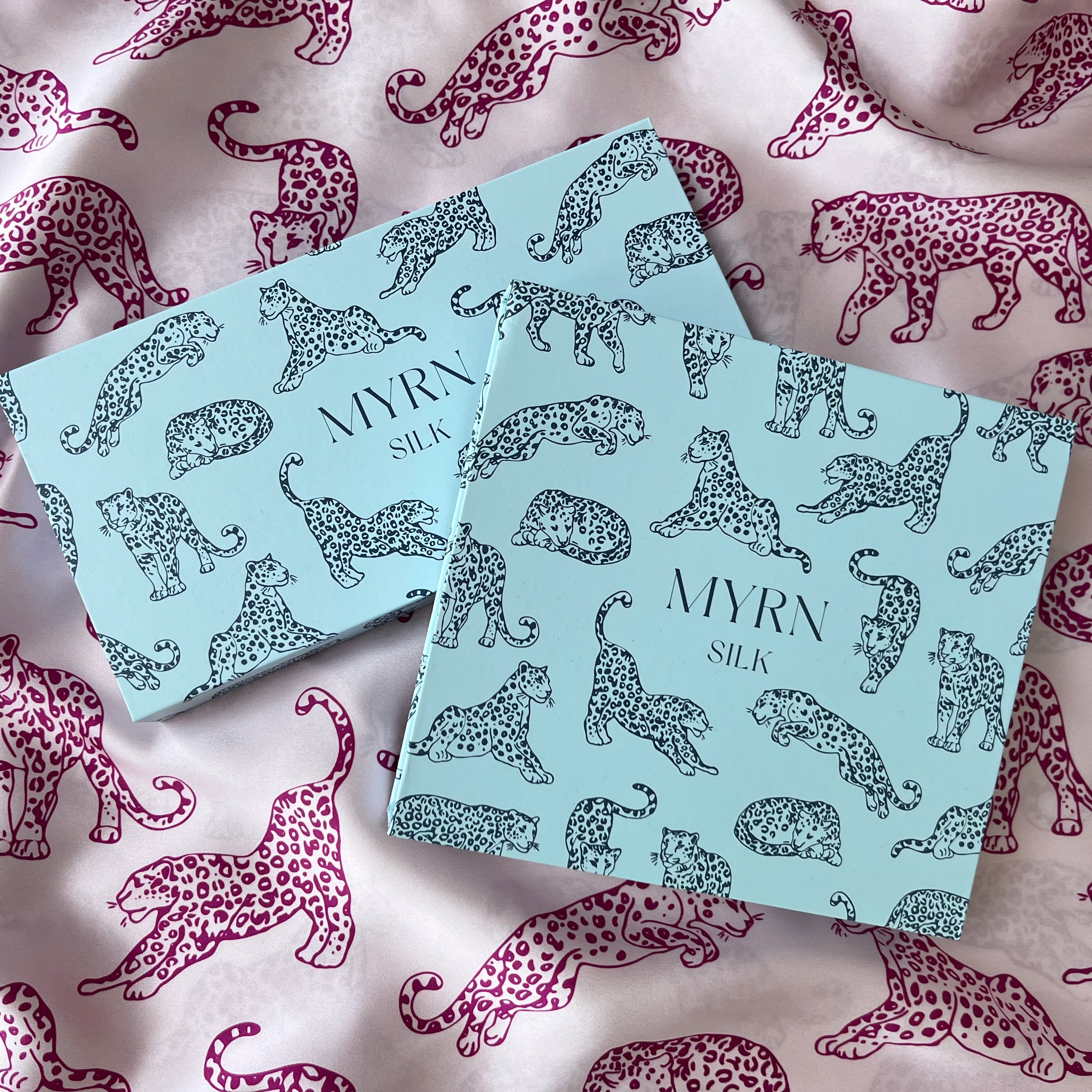 Two MYRN Silk branded presentation boxes laying on draped pink leopard print fabric.