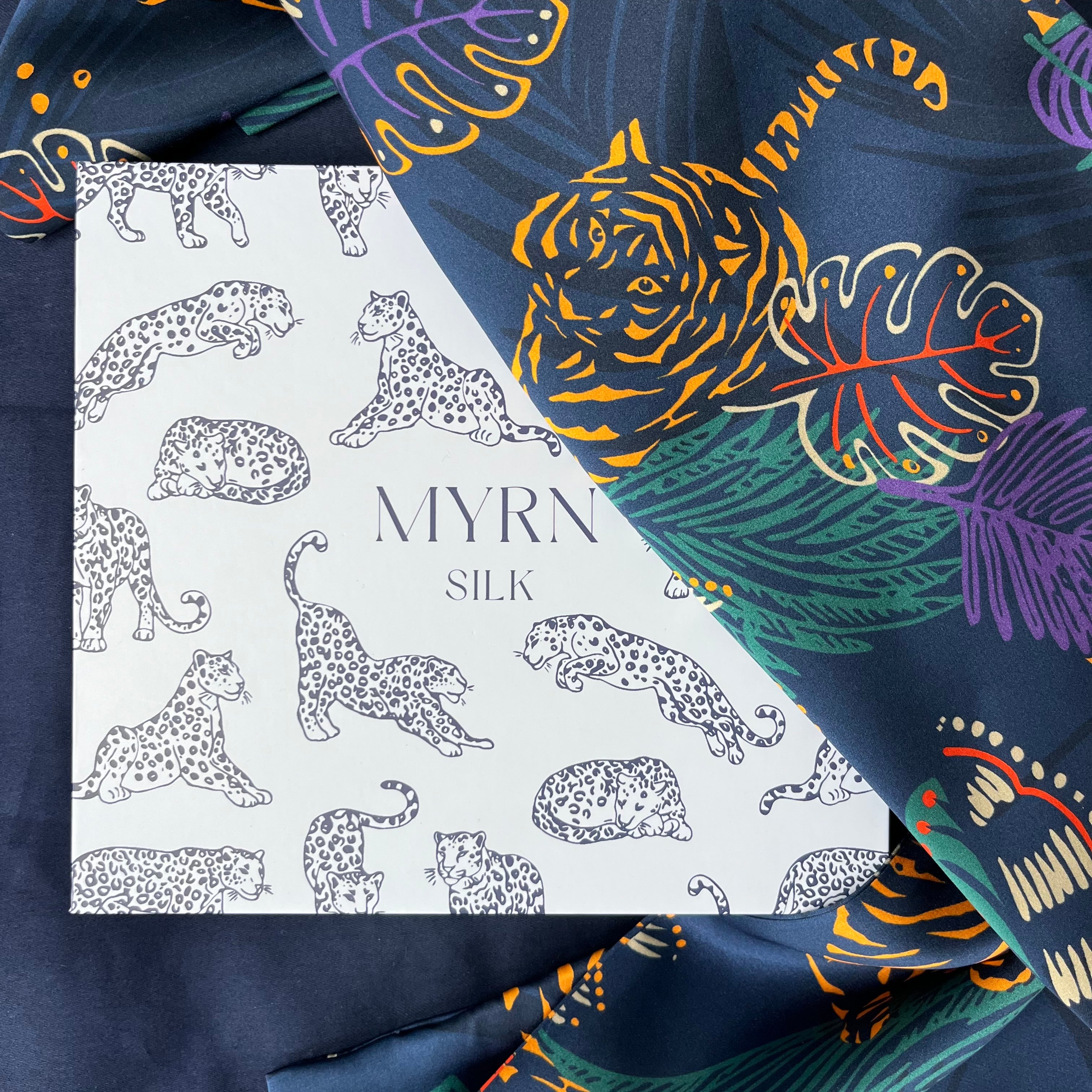 MYRN Silk tiger print pillowcase draped over a MYRN Silk branded leopard print gift box on a dark blue background.