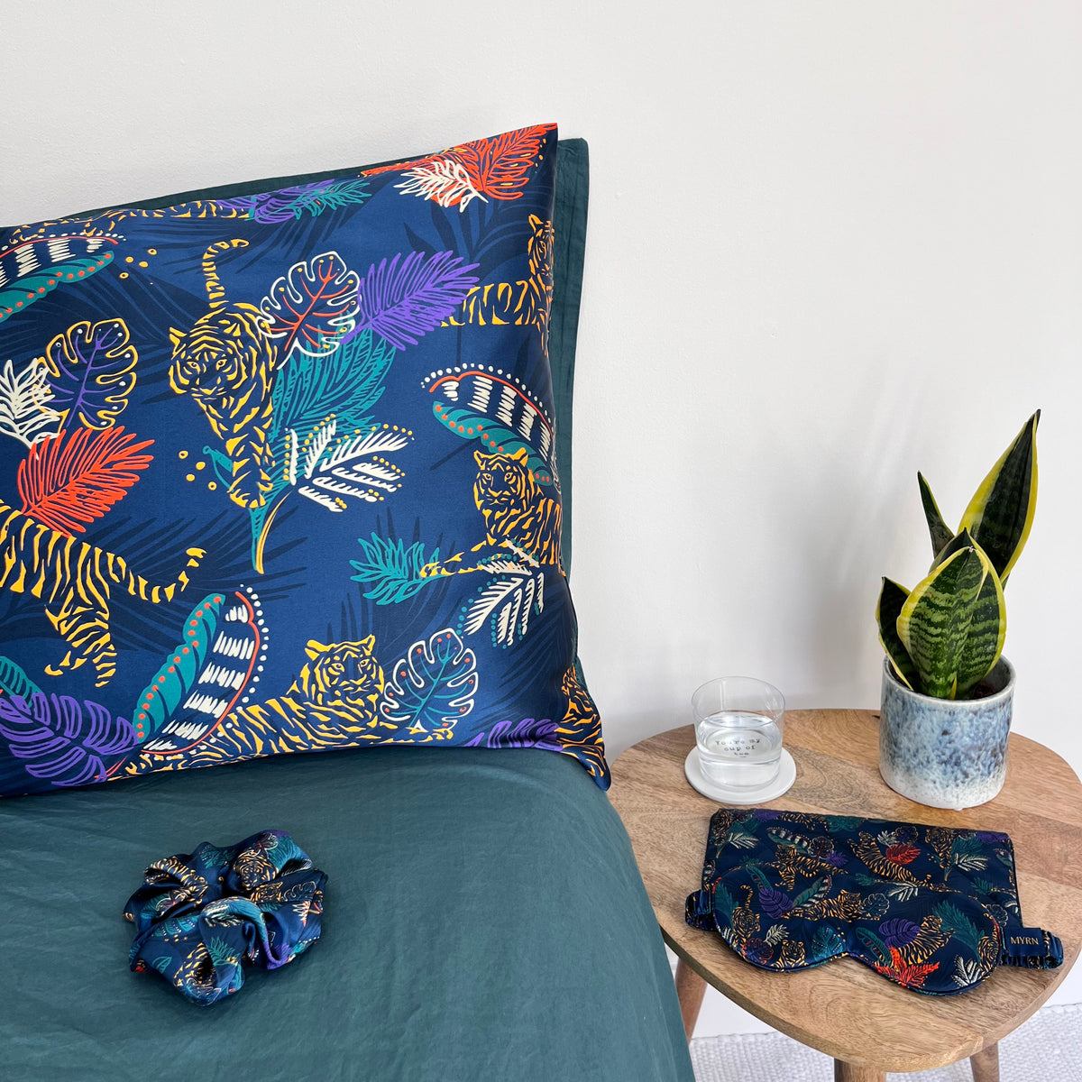 MYRN Silk tiger print pillowcase on a bed with a matching silk scrunchie and a matching silk eye mask with travel case on the side table.